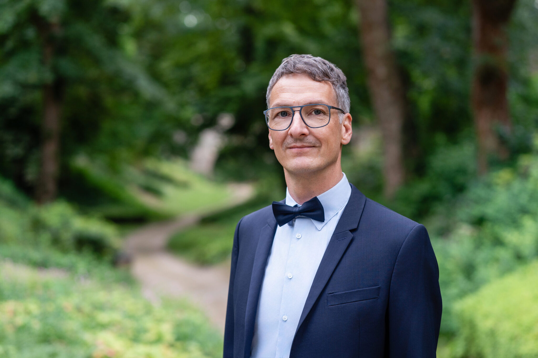Robert Kiefner steht in einem dunkelblauen Anzug mit Fliege auf einem Waldweg